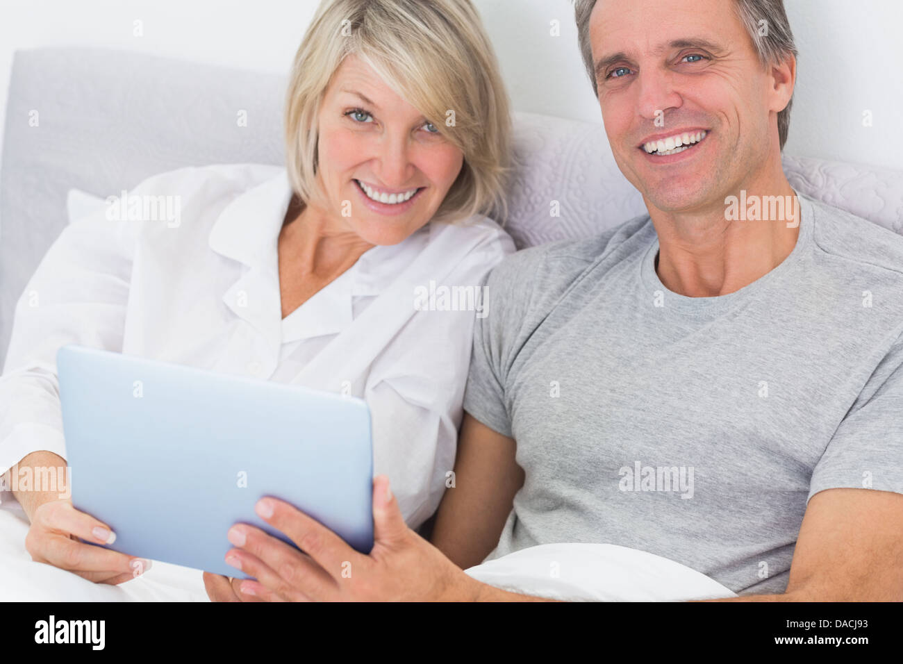 Couple using tablet pc in bed smiling at camera Banque D'Images