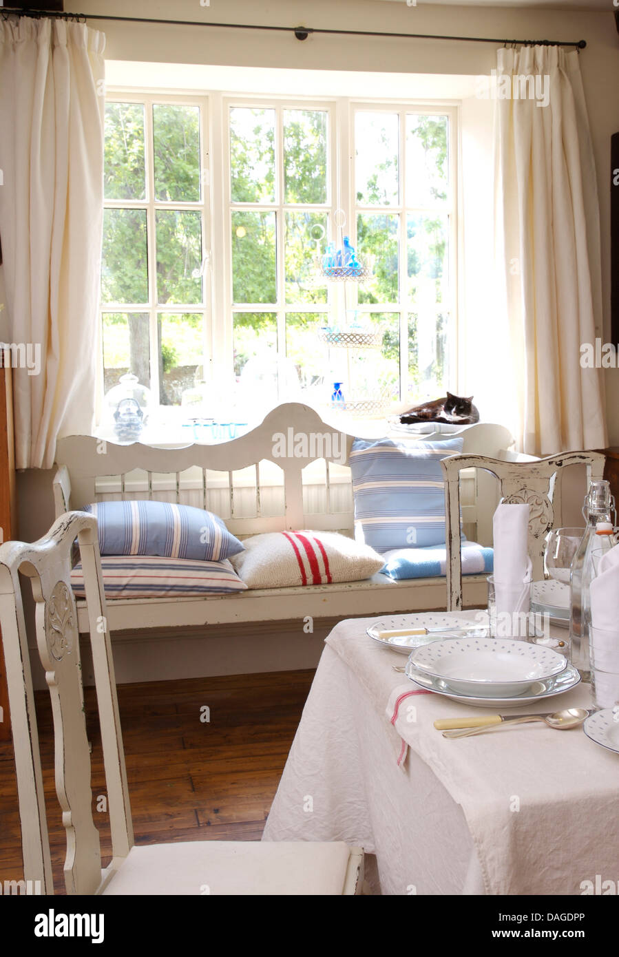 Coussin rayé bleu sur blanc s'installer en face de la fenêtre avec des rideaux de lin blanc dans la salle à manger avec un tissu blanc sur la table Banque D'Images