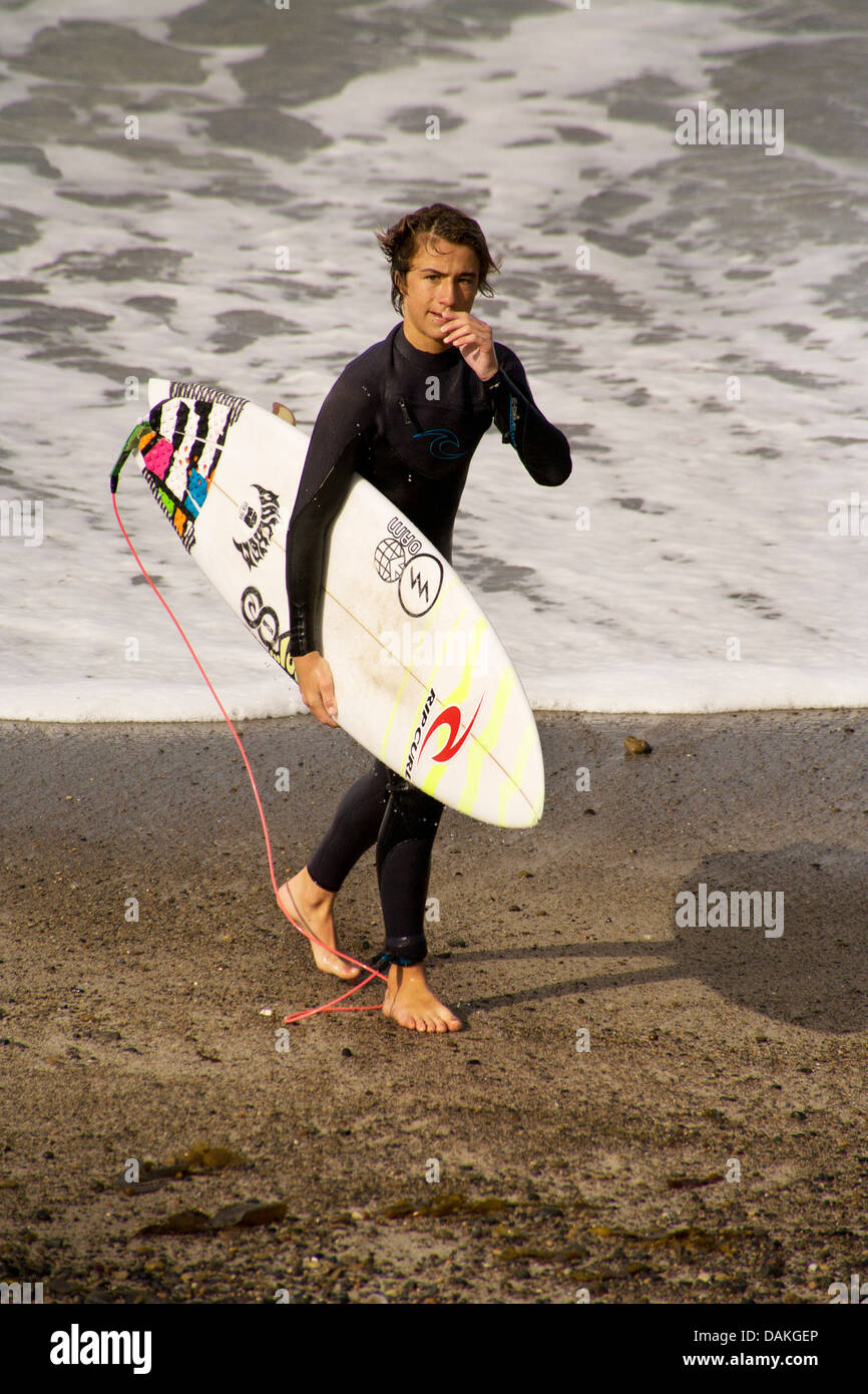 Wearing wetsuit et transportant surfboard, teenage boy membre de l'équipe de surf de l'école secondaire émerge de l'océan Pacifique. Banque D'Images