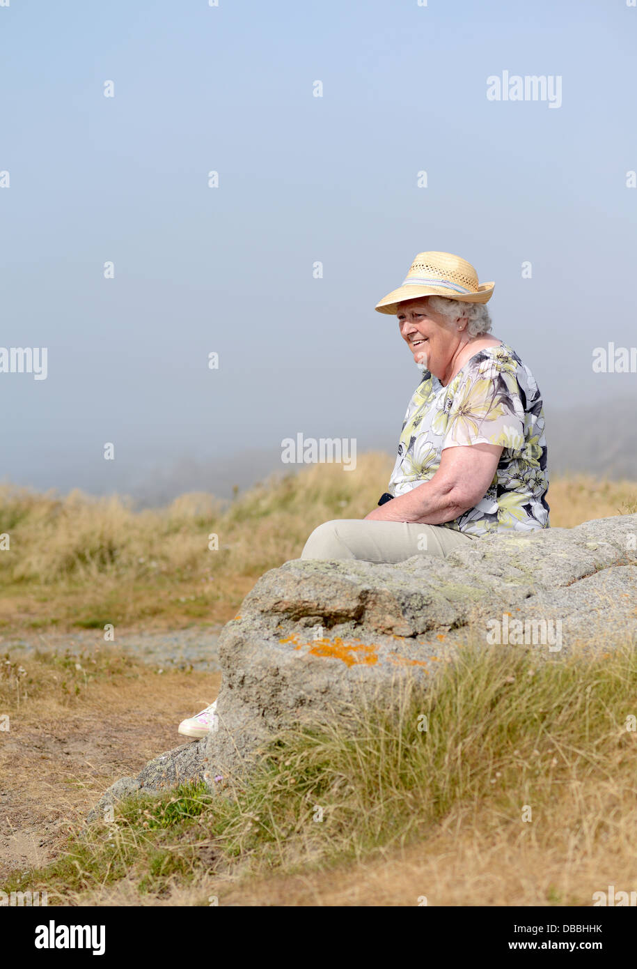 Une vieille dame portant un chapeau de paille assis sur des roches à Guernesey Banque D'Images
