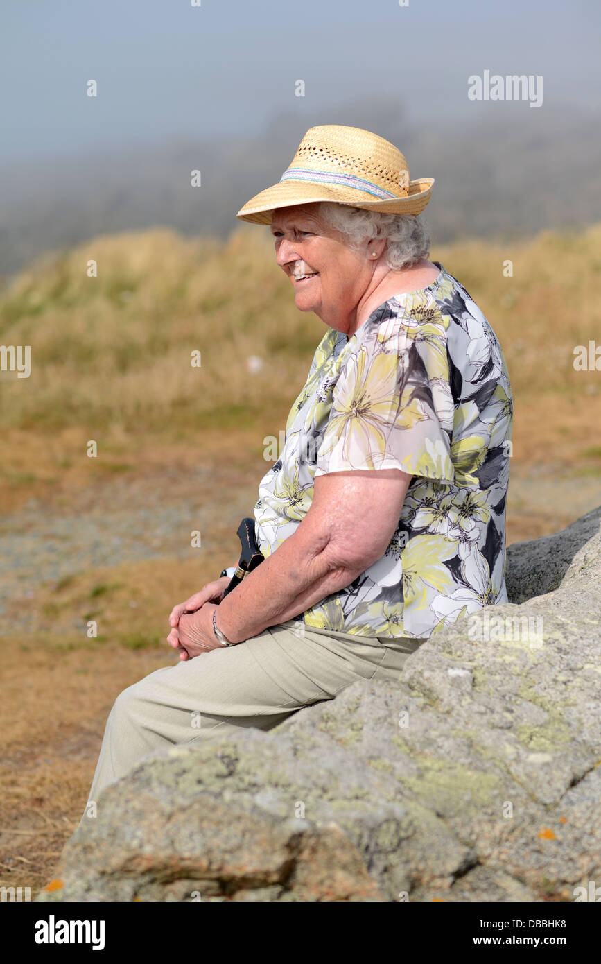 Une vieille dame portant un chapeau de paille assis sur des roches à Guernesey Banque D'Images