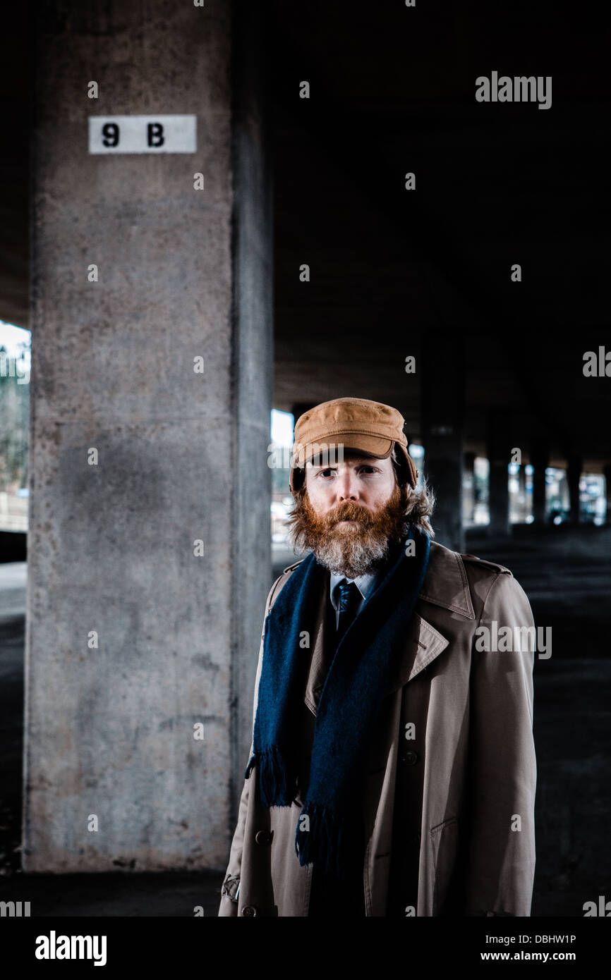 Homme barbu en costume et manteau, bonnet et écharpe debout dans un béton, urbain. Banque D'Images