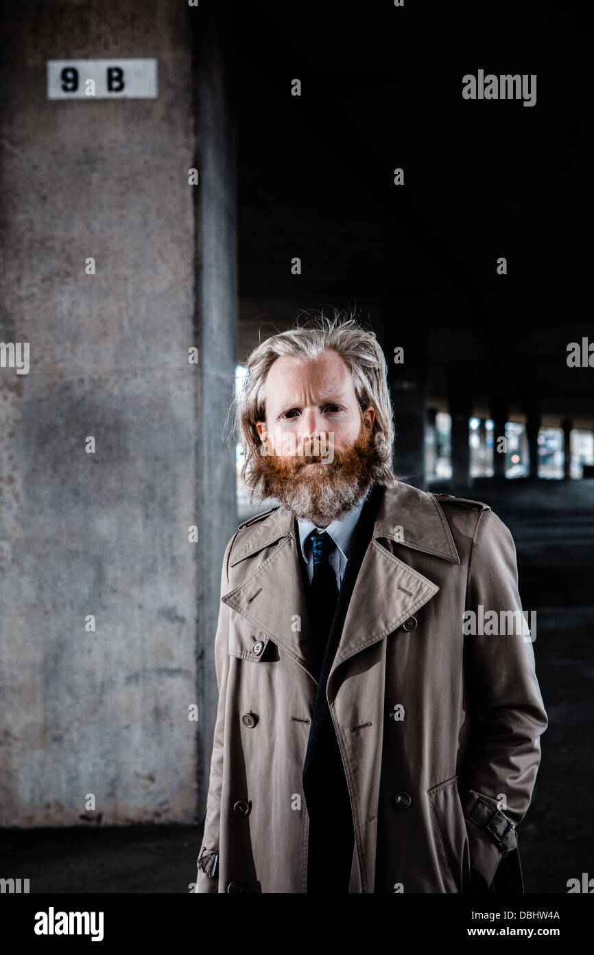 Homme barbu en costume et manteau debout dans un béton, urbain. Banque D'Images