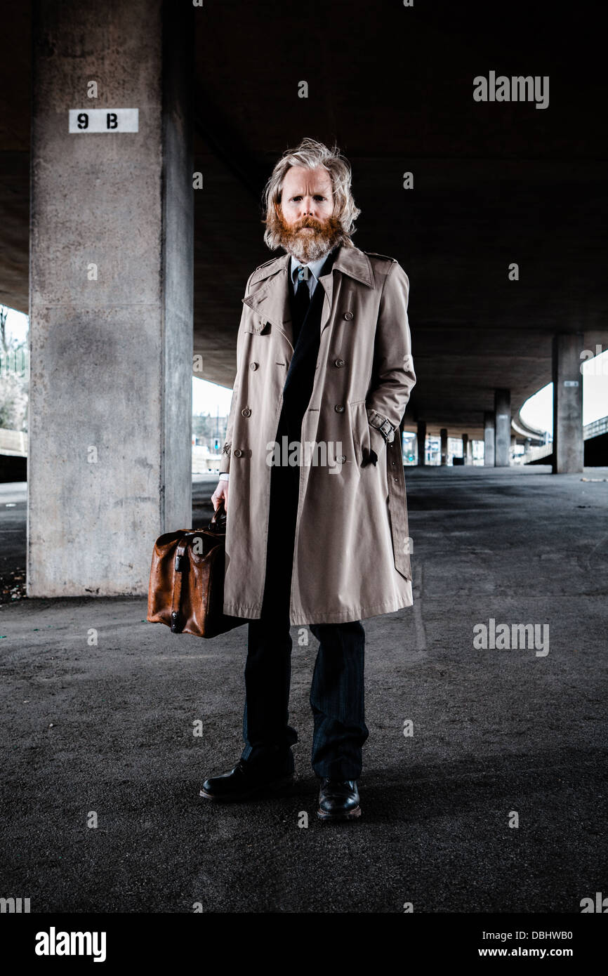 Homme barbu en costume et manteau tenant un vieux cartable en cuir debout dans un béton, urbain. Banque D'Images