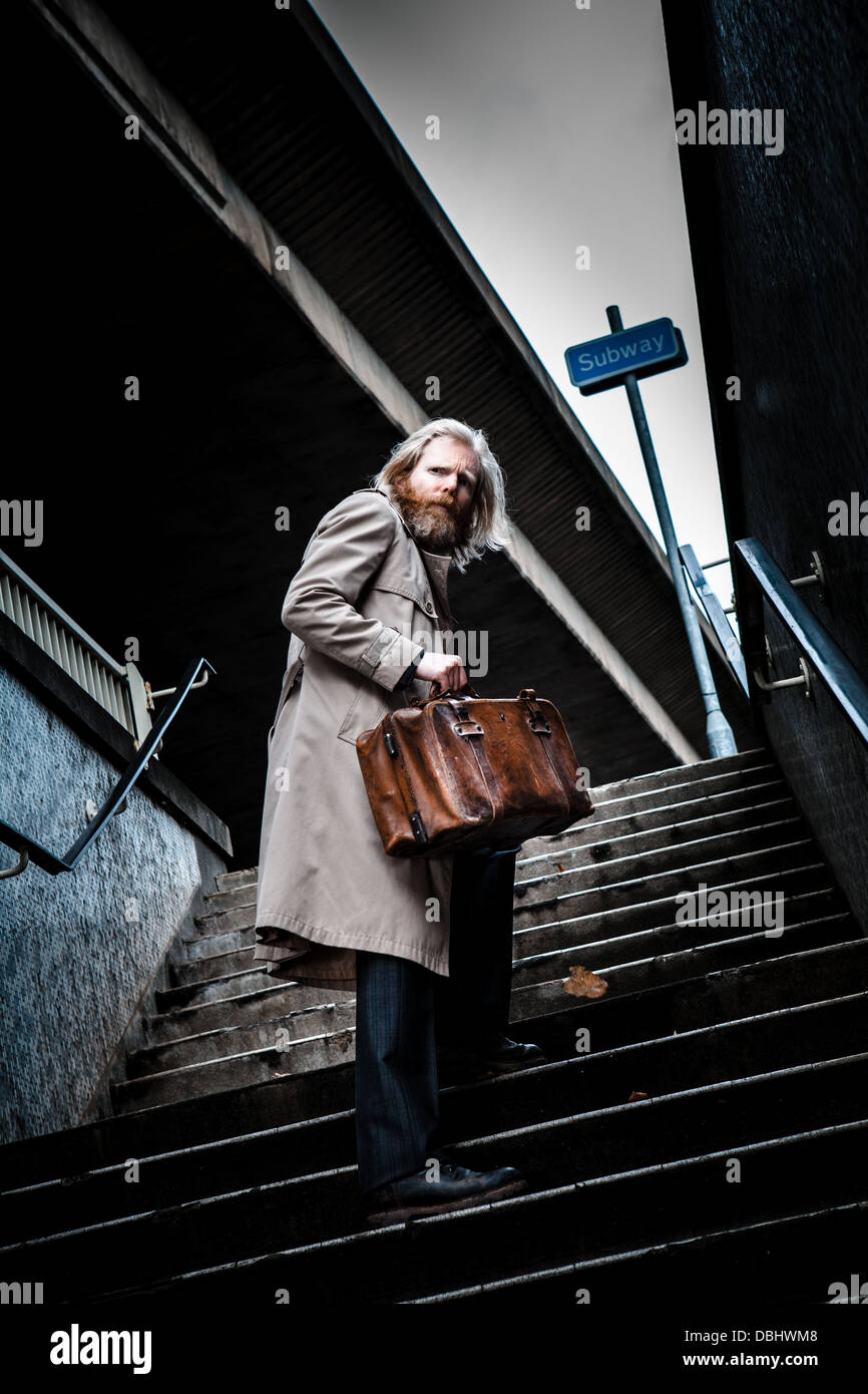 Homme barbu en costume et manteau tenant un vieux cartable en cuir à monter pas hors d'un passage inférieur et à la hâte. Banque D'Images