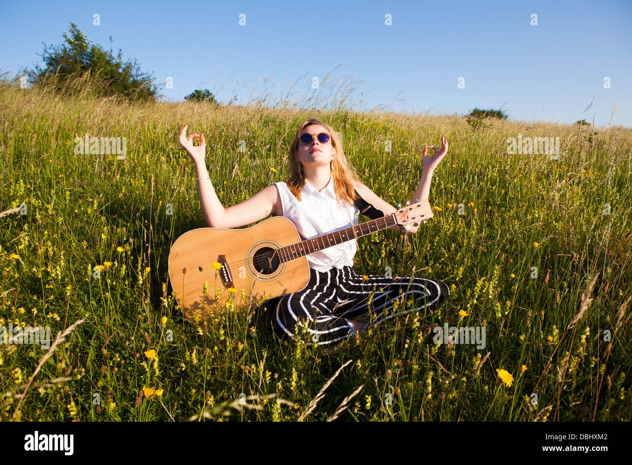 Adolescente portant des lunettes de soleil assis dans une pose de méditation avec une guitare sur ses genoux dans les champs. À l'extérieur. L'été. Banque D'Images