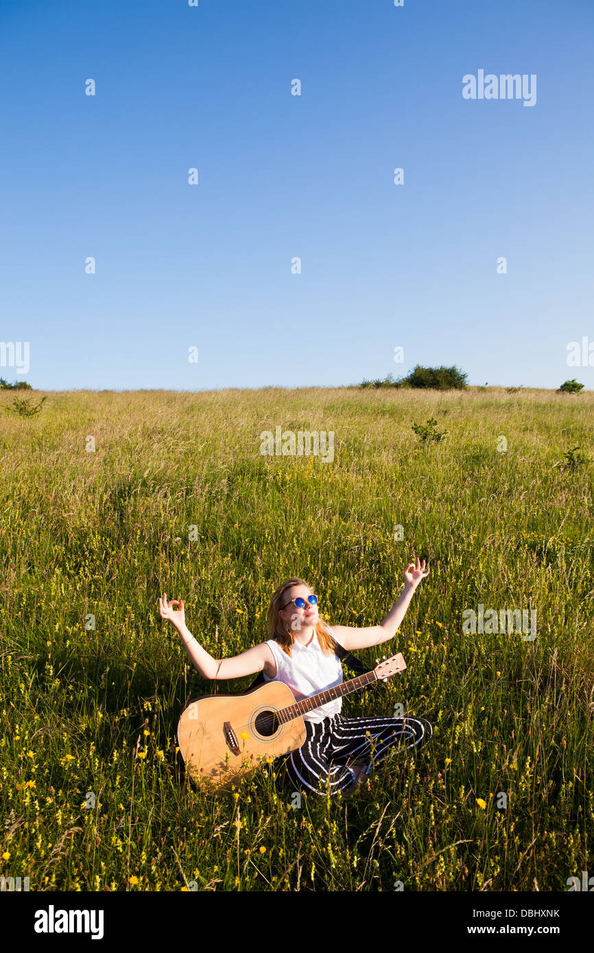 Adolescente portant des lunettes de soleil assis dans une pose de méditation avec une guitare sur ses genoux dans les champs. À l'extérieur. L'été. Banque D'Images