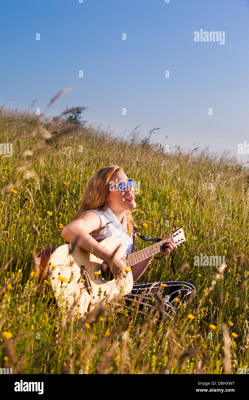 Teenage girl wearing the suglasses assis dans un champ herbeux jouant de la guitare. À l'extérieur. L'été. Banque D'Images