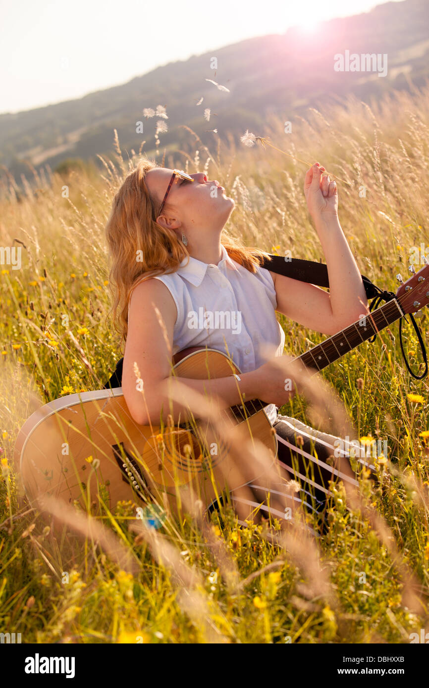 Adolescente portant des lunettes de soleil assis dans un champ d'herbe avec une guitare sur ses genoux qui souffle sur un dandilion tête de semences. À l'extérieur. L'été. Banque D'Images