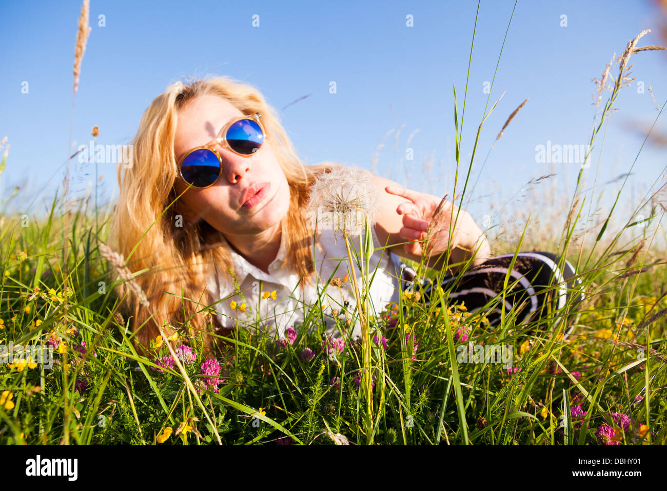 Teenage girl wearing sunglasses assis dans un champ herbeux qui souffle sur un dandilion tête de semences. À l'extérieur. L'été. Banque D'Images