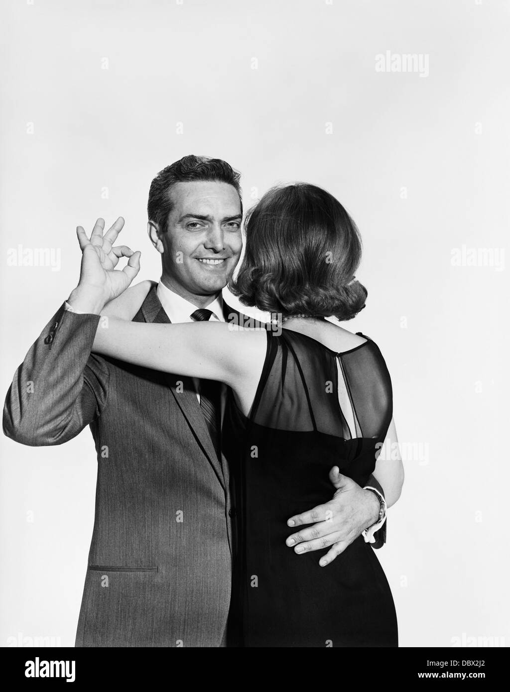 1960 YOUNG MAN LOOKING AT CAMERA FAIRE BON GESTE DERRIÈRE LE DOS DE FEMME Banque D'Images