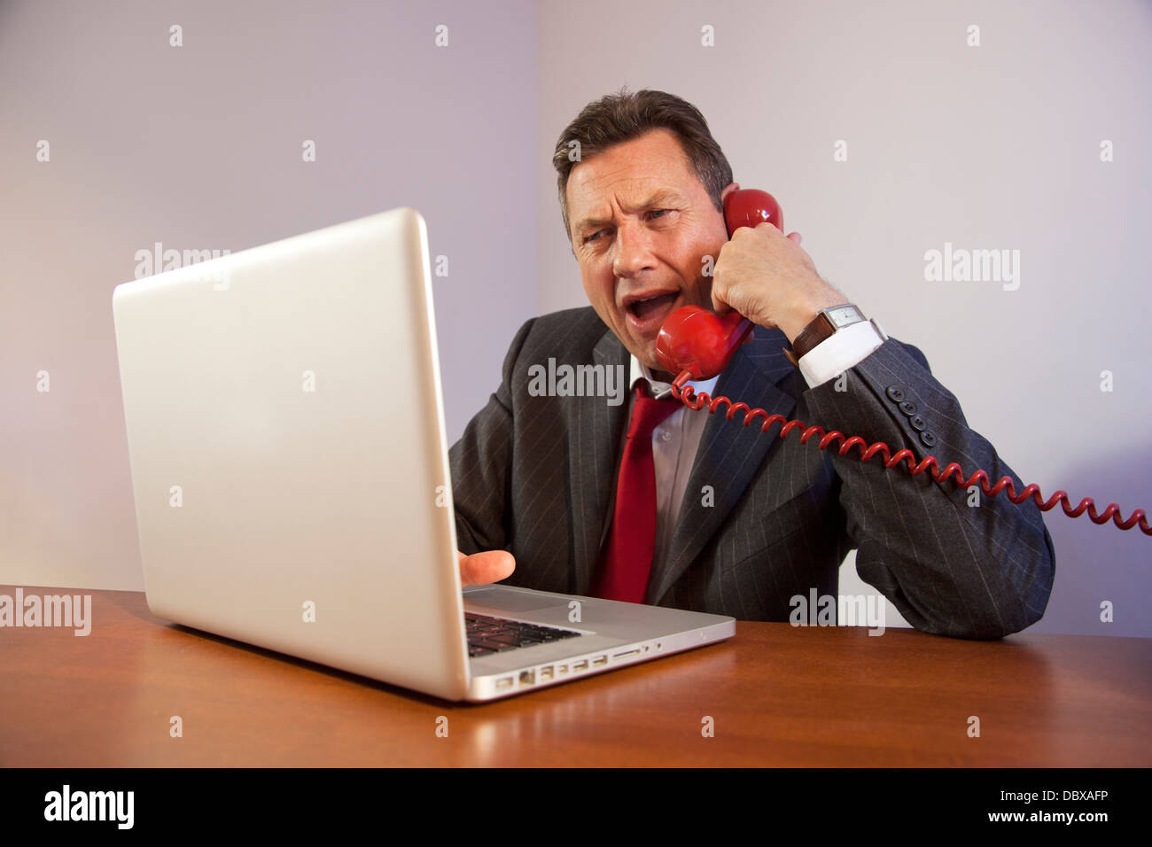 Porter un costume homme en colère, criant vers le bas un téléphone rouge  alors qu'il était assis en face d'un ordinateur portable sur un bureau  Photo Stock - Alamy