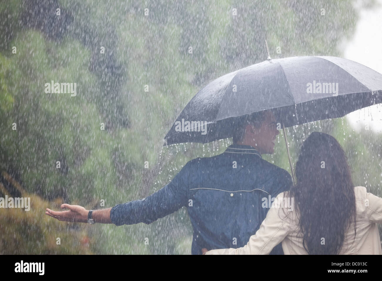 Couple walking with arms outstretched under umbrella in rain Banque D'Images