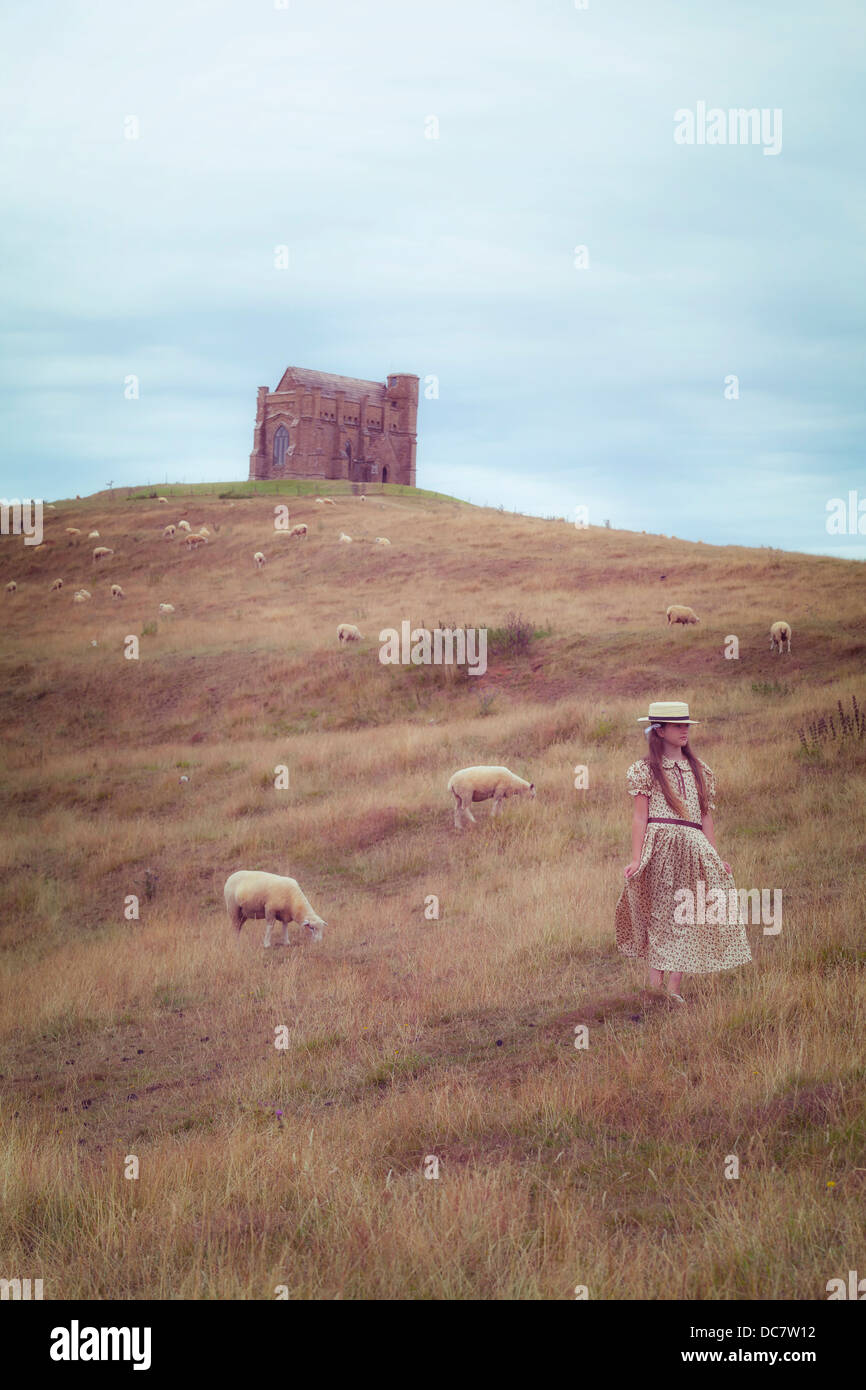 Une fille dans une robe vintage sur une prairie avec moutons, dans l'arrière-plan d'une chapelle Banque D'Images