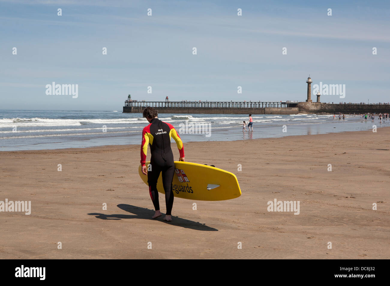 Un sauveteur RNLI en travers de la plage de sable vers la mer avec une planche de surf sous le bras, Whitby, dans le Yorkshire. Banque D'Images