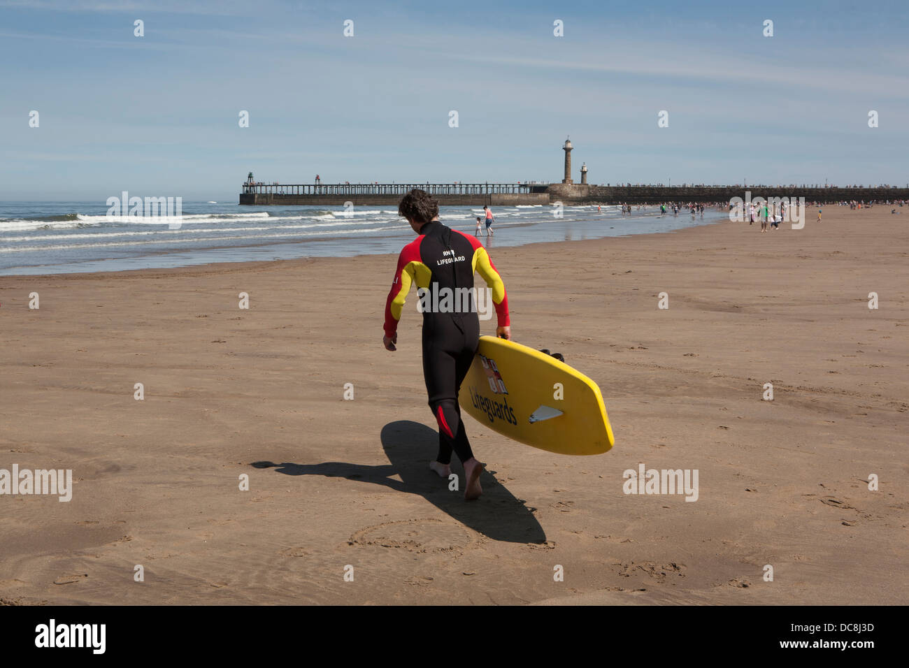 Un sauveteur RNLI en travers de la plage de sable vers la mer avec une planche de surf sous le bras, Whitby, dans le Yorkshire. Banque D'Images