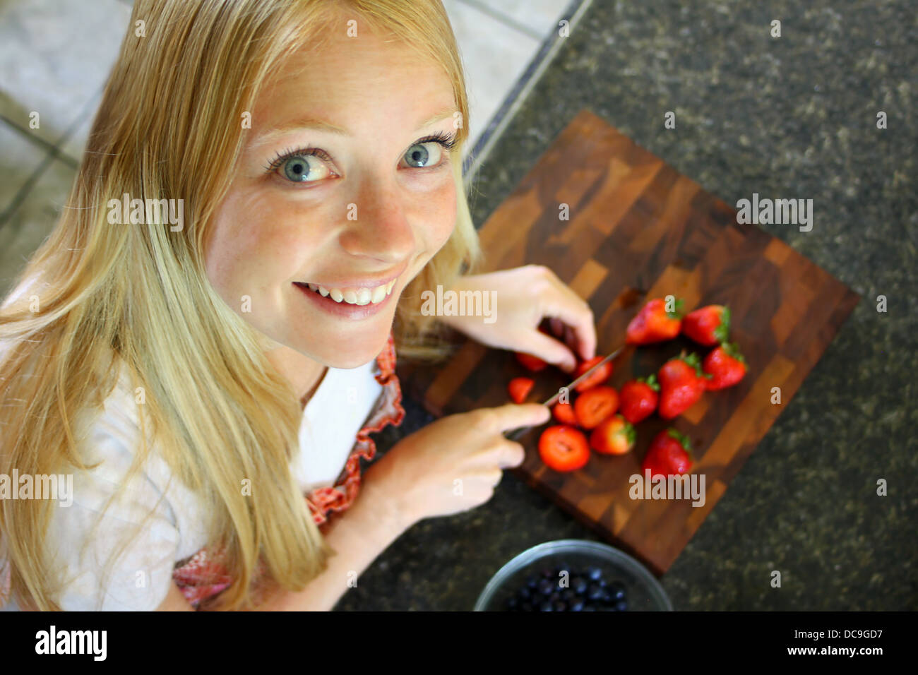 Une jolie femme est à la recherche jusqu'à l'appareil photo en souriant, alors qu'elle coupe les fraises. Banque D'Images