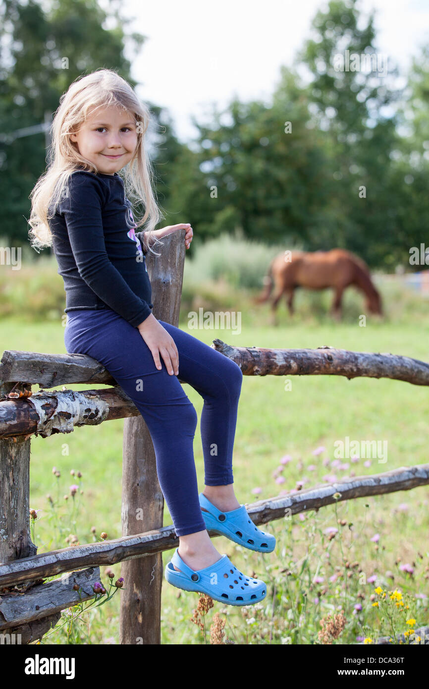 Petit pays girl sitting on fence avec les chevaux sur contexte en village à jour d'été. Peuple russe dans les régions rurales de la Russie Banque D'Images