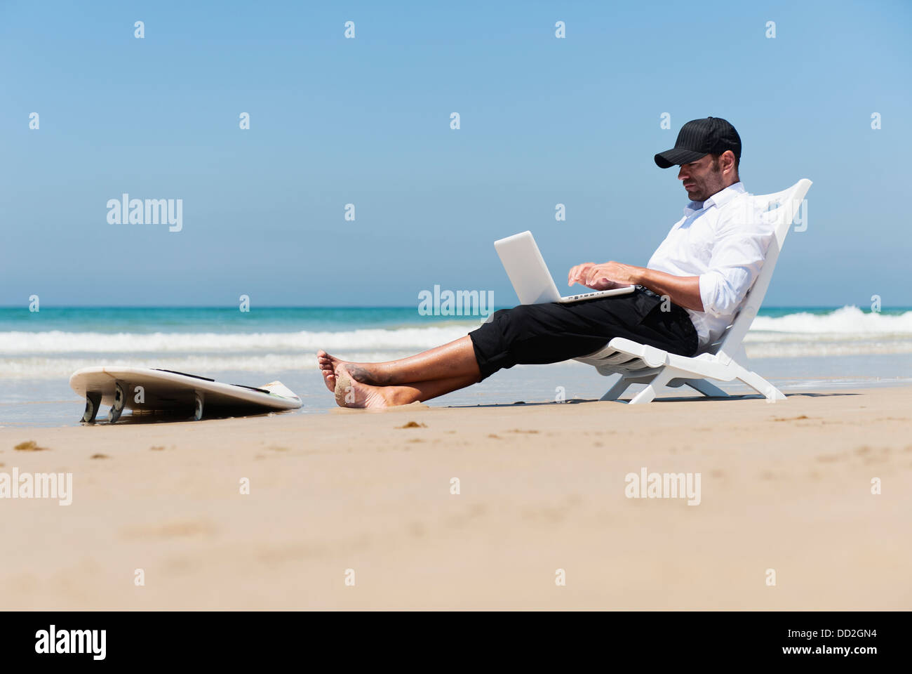 Un homme est assis sur une chaise de plage sur la plage de travailler sur un ordinateur portable avec sa séance de surf à ses pieds ; l'Espagne Banque D'Images
