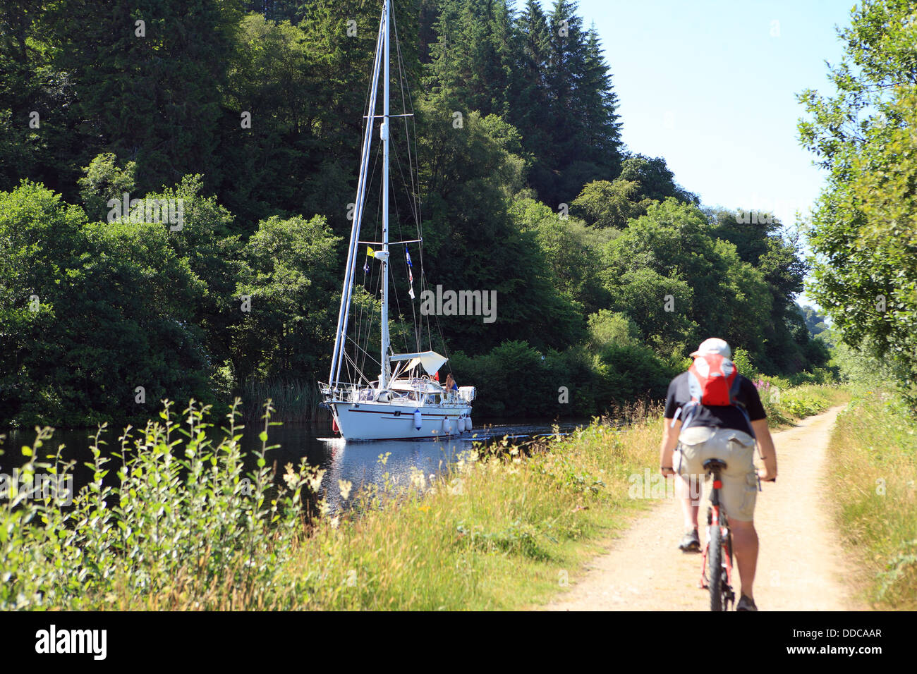 Cycliste sur la piste cyclable du Canal Crinan passant un yacht dans le canal à Argyll en Écosse Banque D'Images