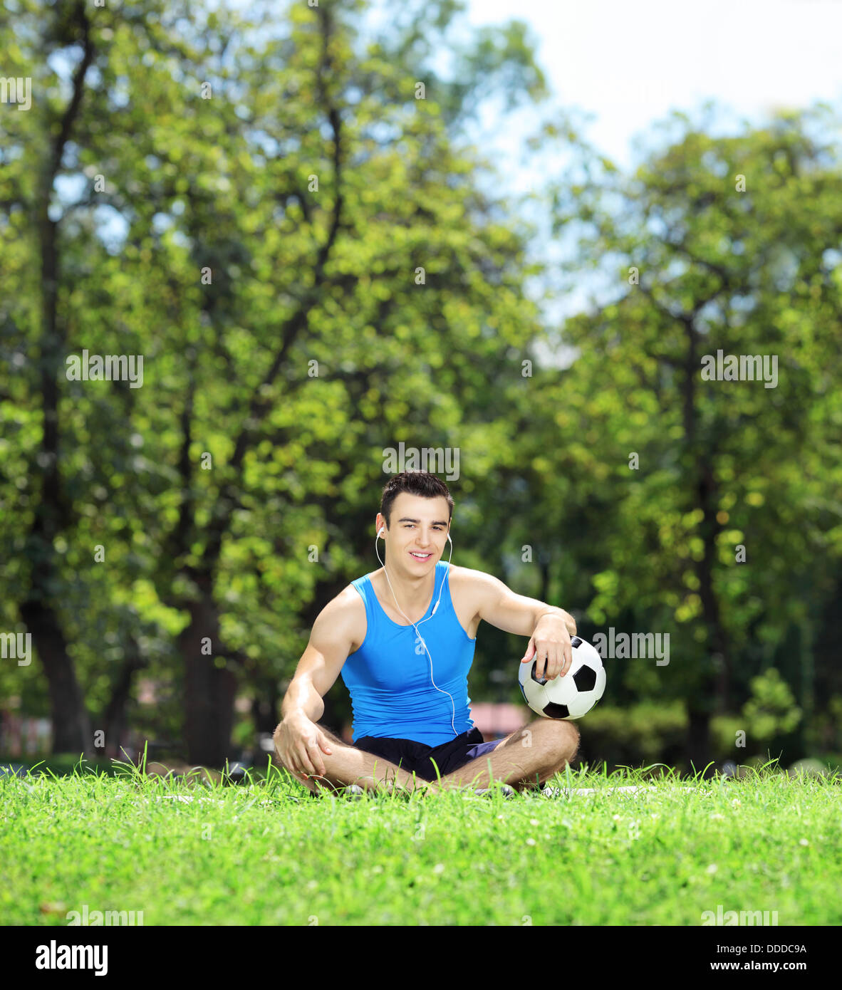 Jeune athlète masculin souriant assis sur l'herbe verte avec un ballon de soccer dans un parc Banque D'Images