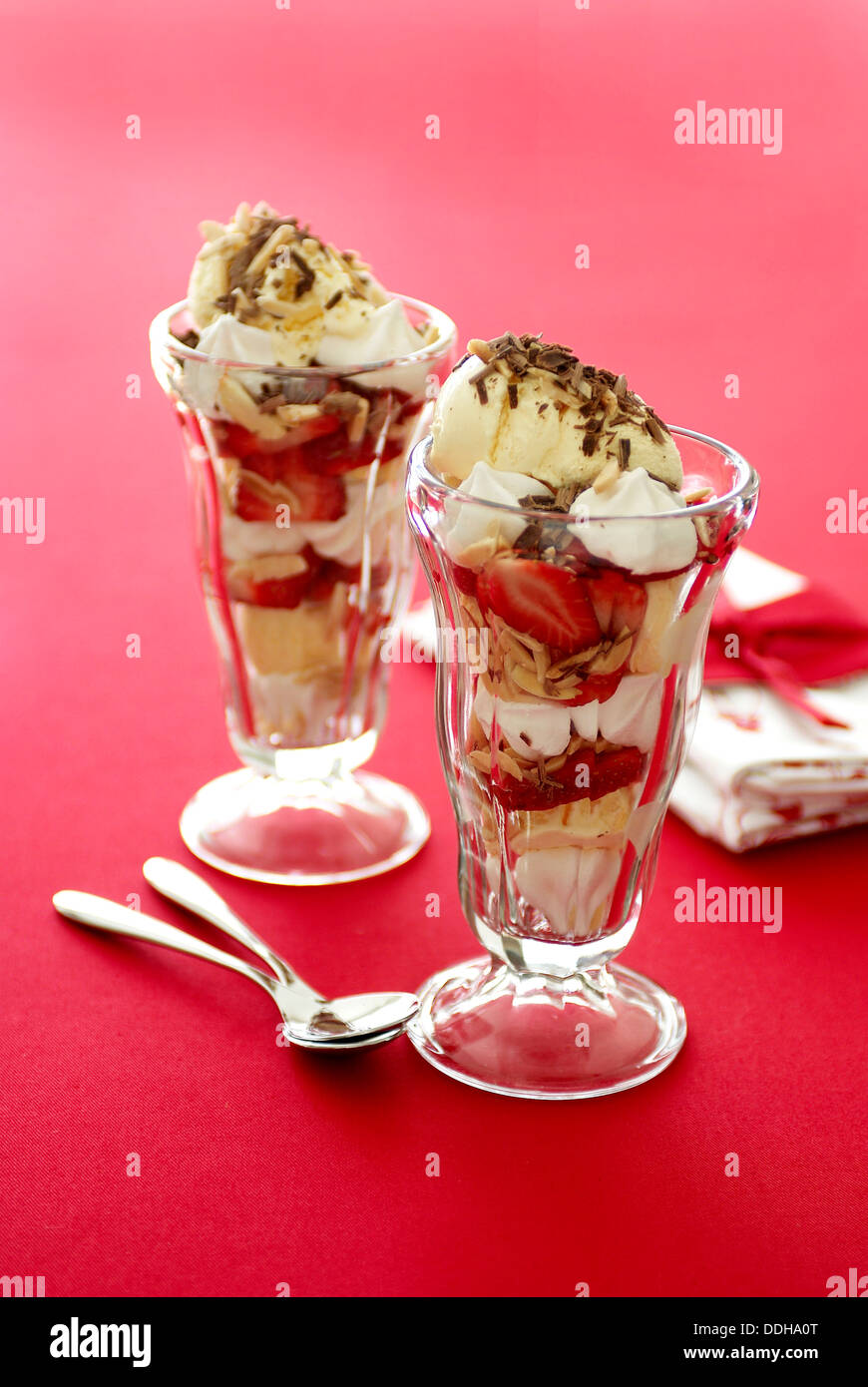 Deux coupes de fraises à la crème, meringues, crème glacée et chocolat sur une surface de table rouge. Banque D'Images