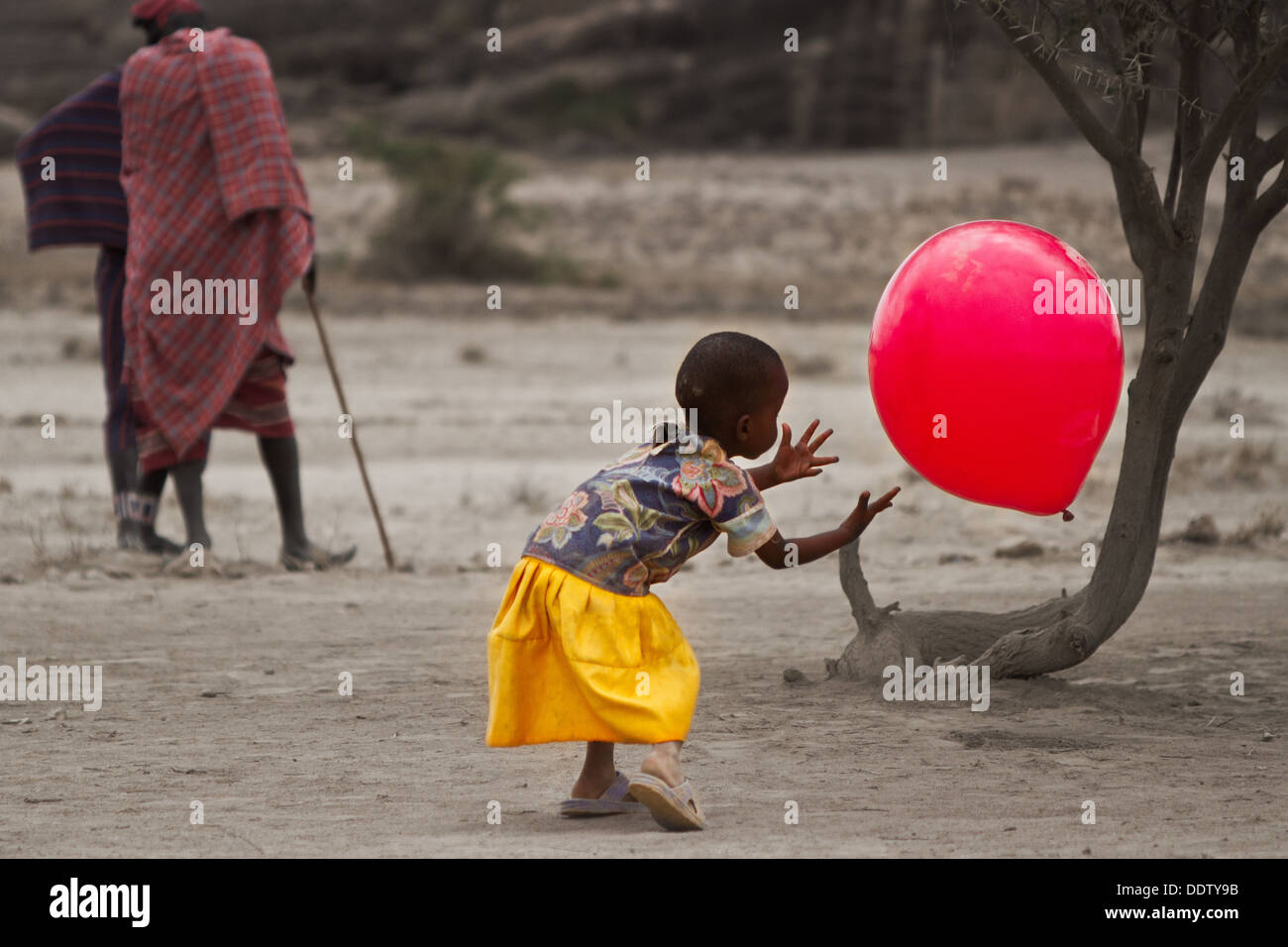 Petite fille jouant à l'extérieur avec de gros ballon rouge, encadré par  tronc de l'arbre, tandis que deux hommes Masai parler en arrière-plan, à la  fois habillé Photo Stock - Alamy