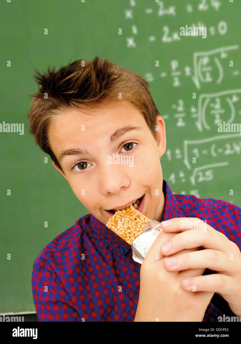 Portrait, écolier, adolescent mordre dans une barre de muesli devant une école tableau avec une équation mathématique Banque D'Images