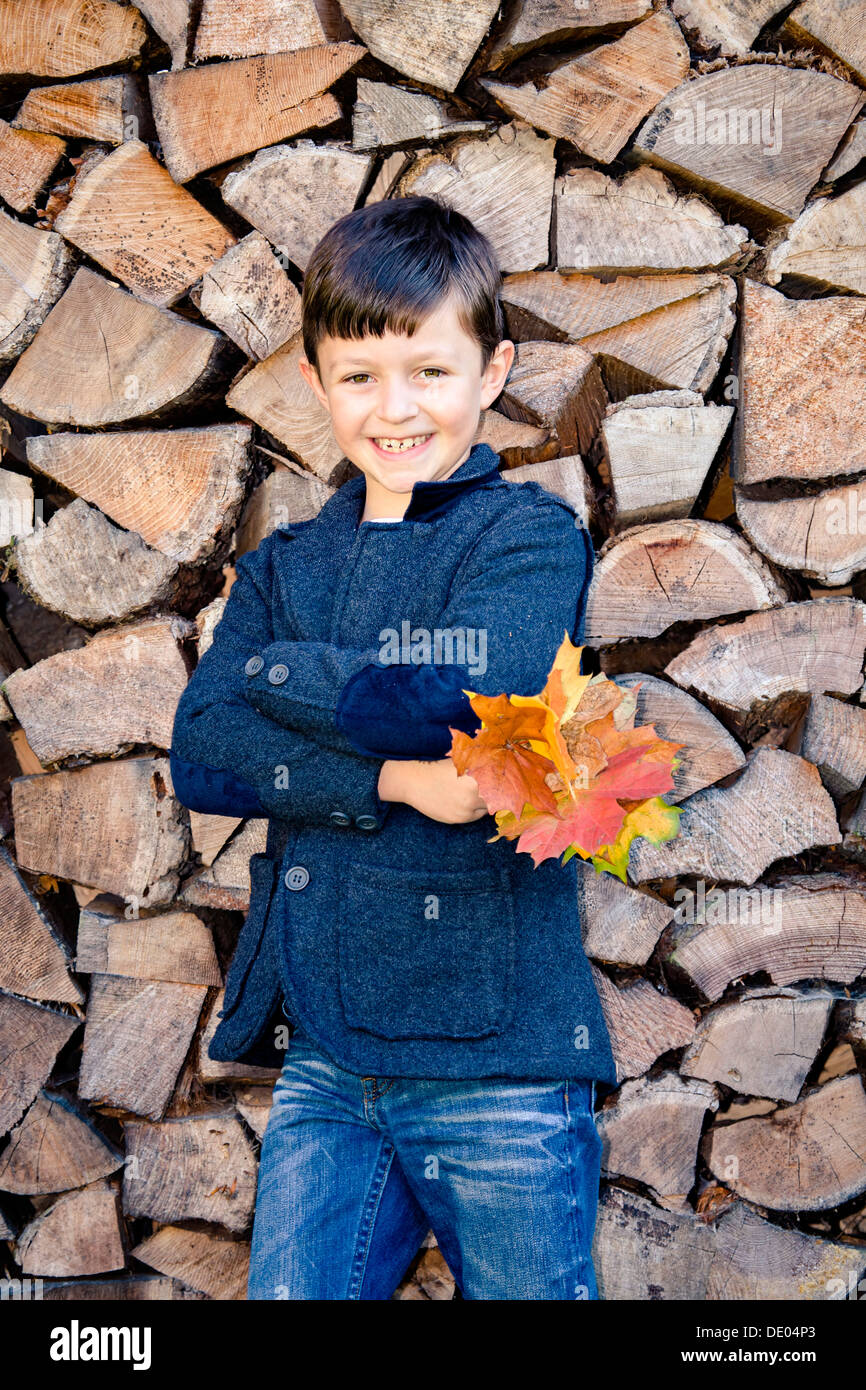 Boy holding Autumn Leaves, debout devant des bois empilés Banque D'Images
