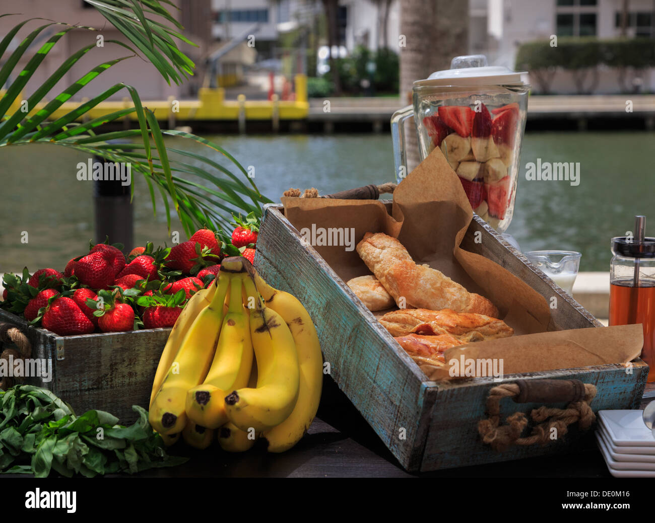Milieu urbain, petit déjeuner au bord de l'eau. Blender avec des morceaux de fruits sur table avec fort de produits de boulangerie, les fraises et les bananes. Banque D'Images