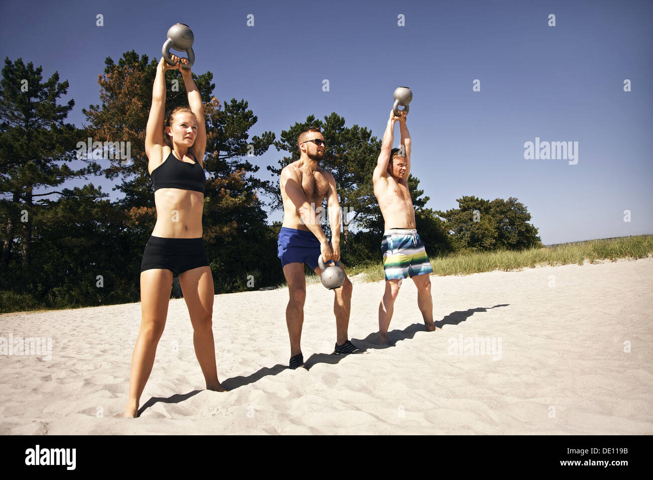 Un groupe d'athlètes balançant une cloche électrique au-dessus de leur tête, sur la plage. Des jeunes faisant l'entraînement Crossfit sur une chaude journée d'été. Banque D'Images