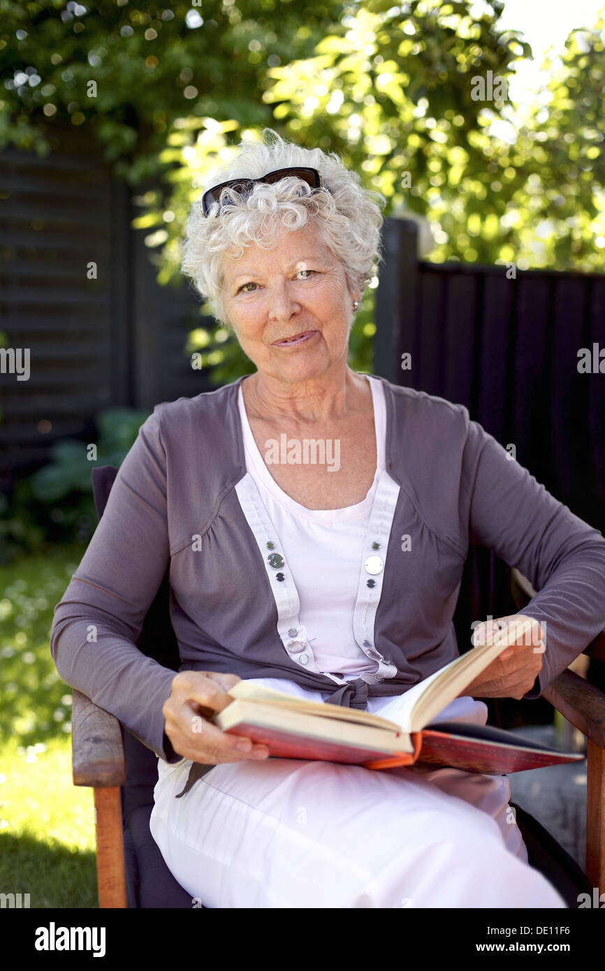 Mature femme assise sur une chaise de jardin avec un livre à la caméra en souriant - Elder couple outdoors Banque D'Images