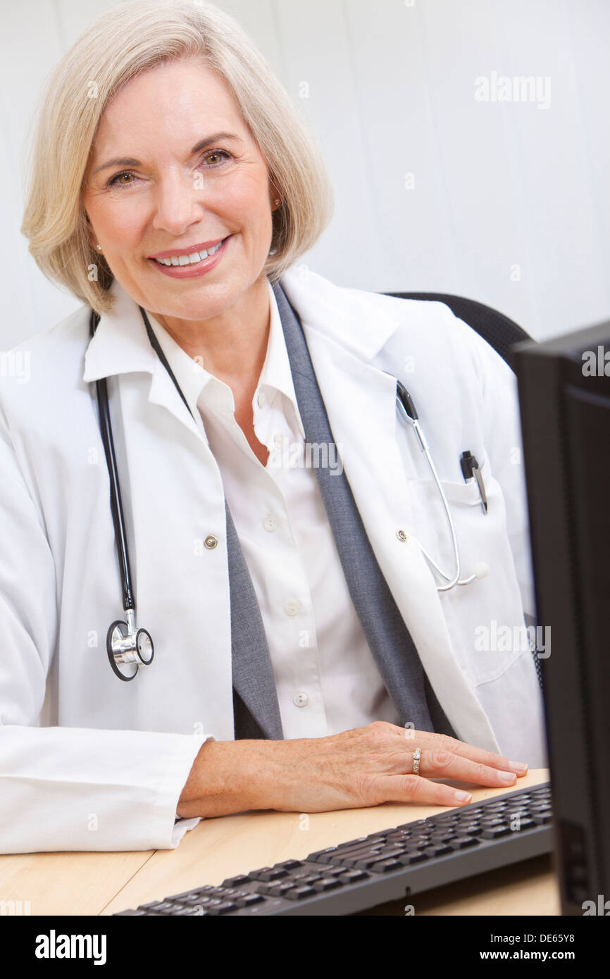 Une médecin assis à un bureau dans un bureau avec un ordinateur, le port d'une blouse blanche et stéthoscope Banque D'Images