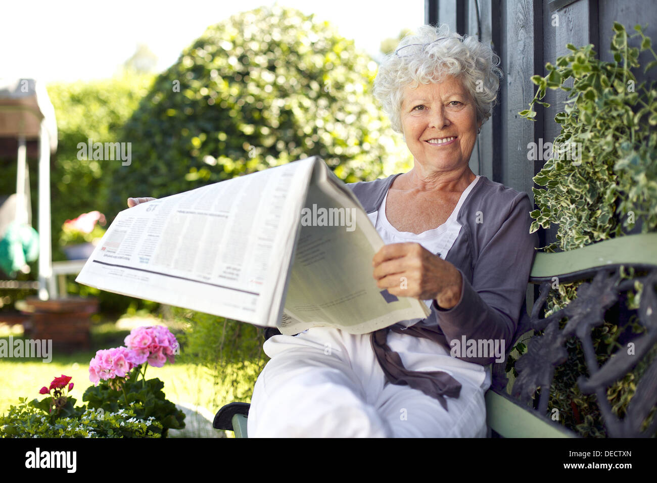 La haute atmosphère femme assise sur un banc dans le jardin lire un journal looking at camera and smiling Banque D'Images