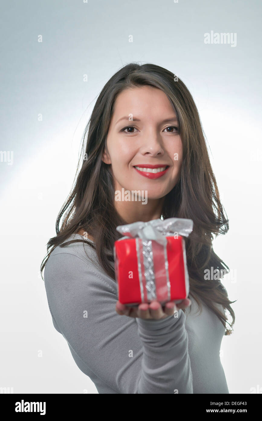 Jolie jeune femme aux longs cheveux brun et un beau sourire doux qui fait un cadeau rouge coloré pour un être cher Banque D'Images