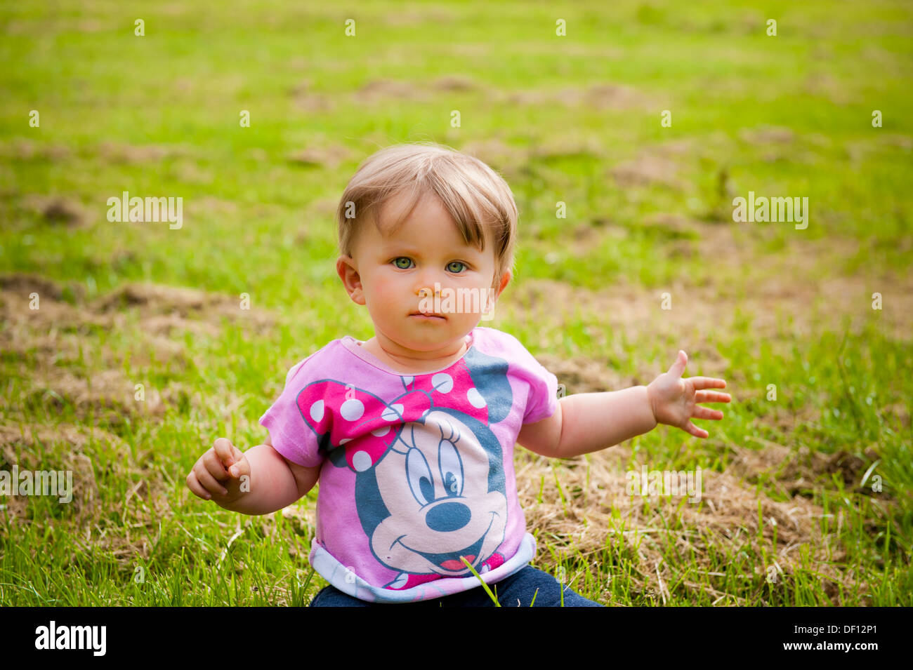 Baby Girl holding arms out assis sur l'herbe avec expression visage grave Banque D'Images