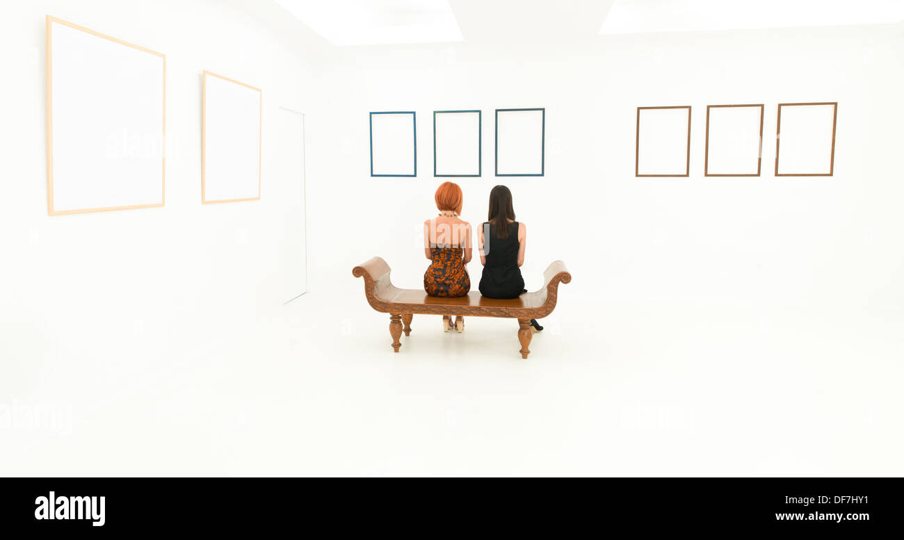 Vue arrière des deux femmes assises sur un banc en bois à vide à images affichées sur un mur en face d'eux Banque D'Images