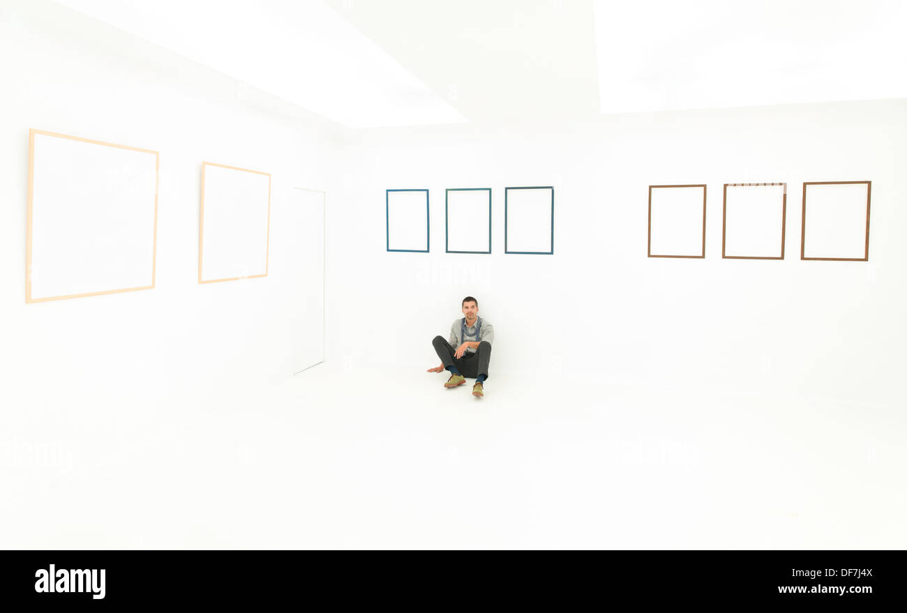 Homme assis sur le sol dans une position de détente, dans une pièce blanche avec des images affichées sur les murs vides Banque D'Images