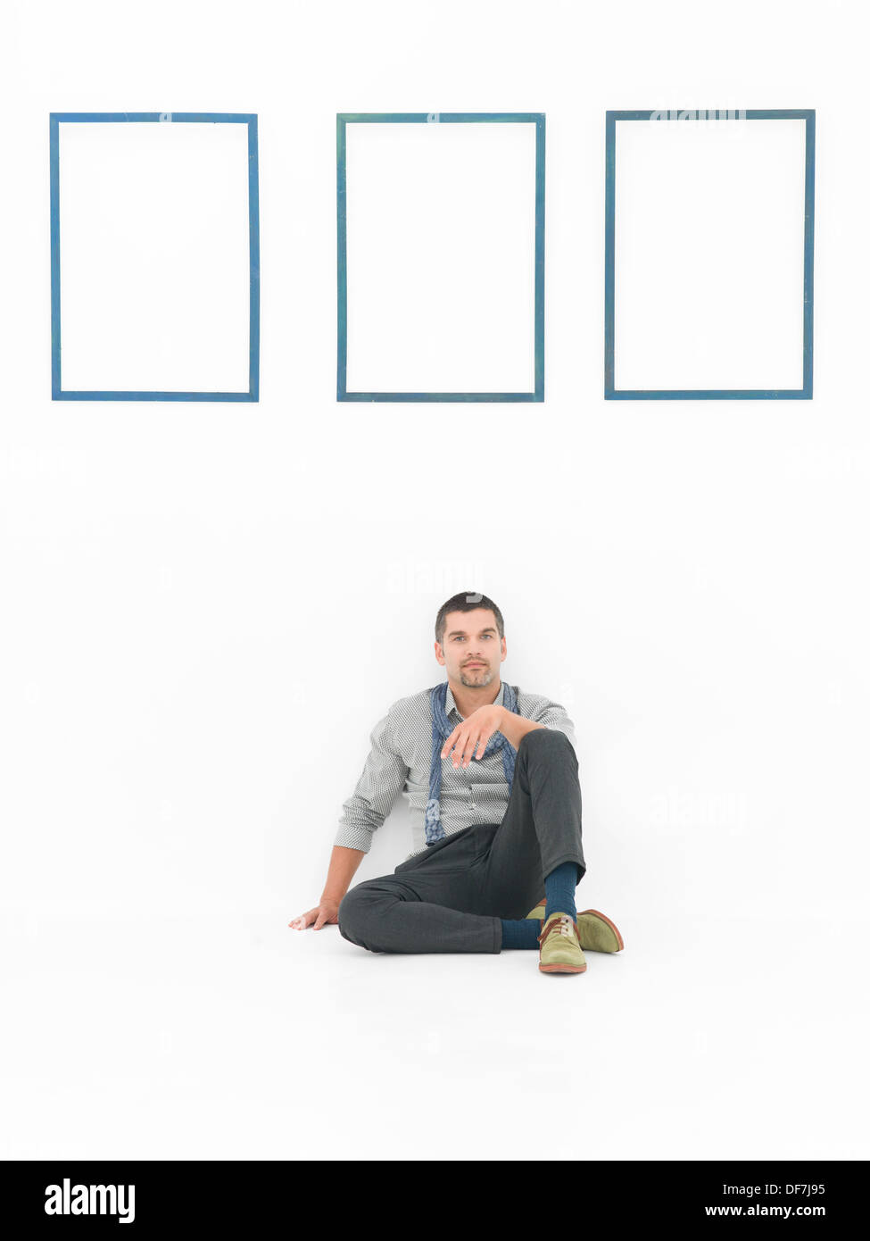 Vue de face de l'homme assis sur le sol dans une posture détendue contre un mur blanc avec trois cadres bleus vides Banque D'Images