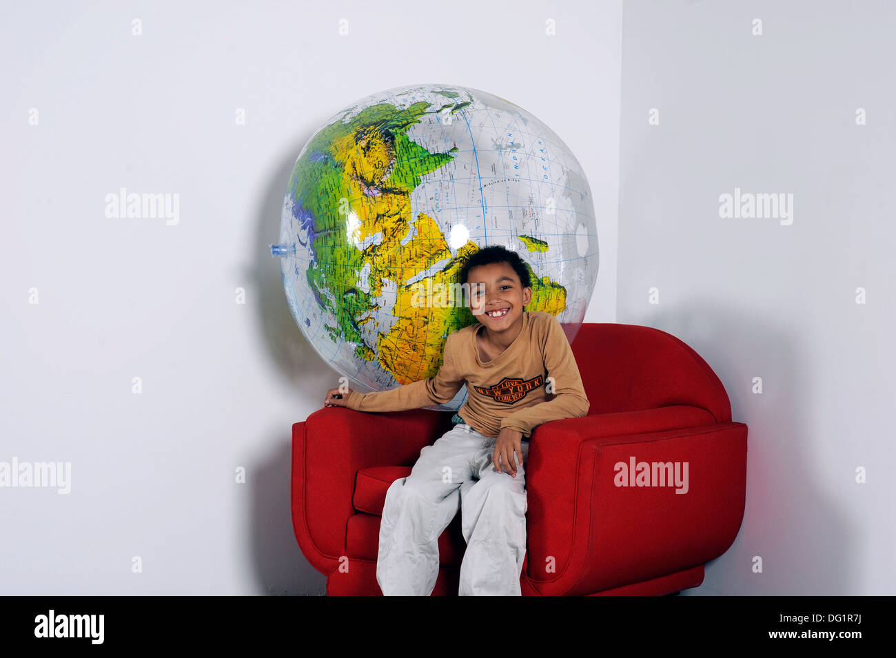Jeune garçon souriant, assis avec un gros ballon globe derrière lui dans un fauteuil rouge Banque D'Images