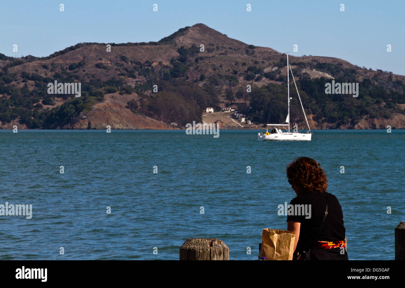 Femme assise à côté de la baie de manger le déjeuner à Sausalito, Californie avec voilier et de l'Angel Island dans l'arrière-plan Banque D'Images