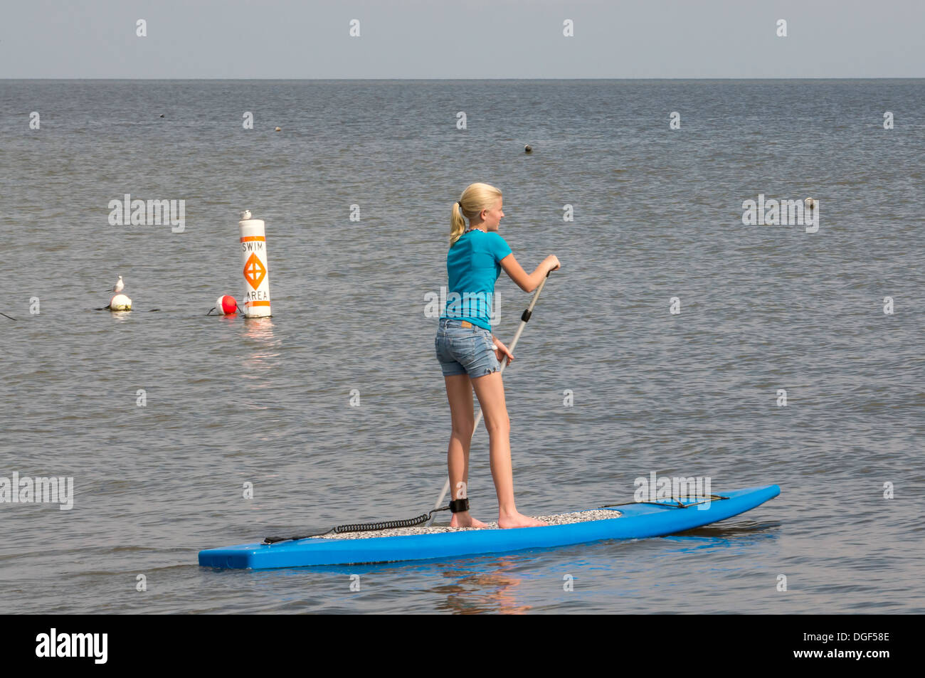 Young woman wearing blue shorts et T-shirt sur le SUP stand up paddleboard sur eaux calmes tout en étant à proximité d'une plage de sable blanc. Banque D'Images