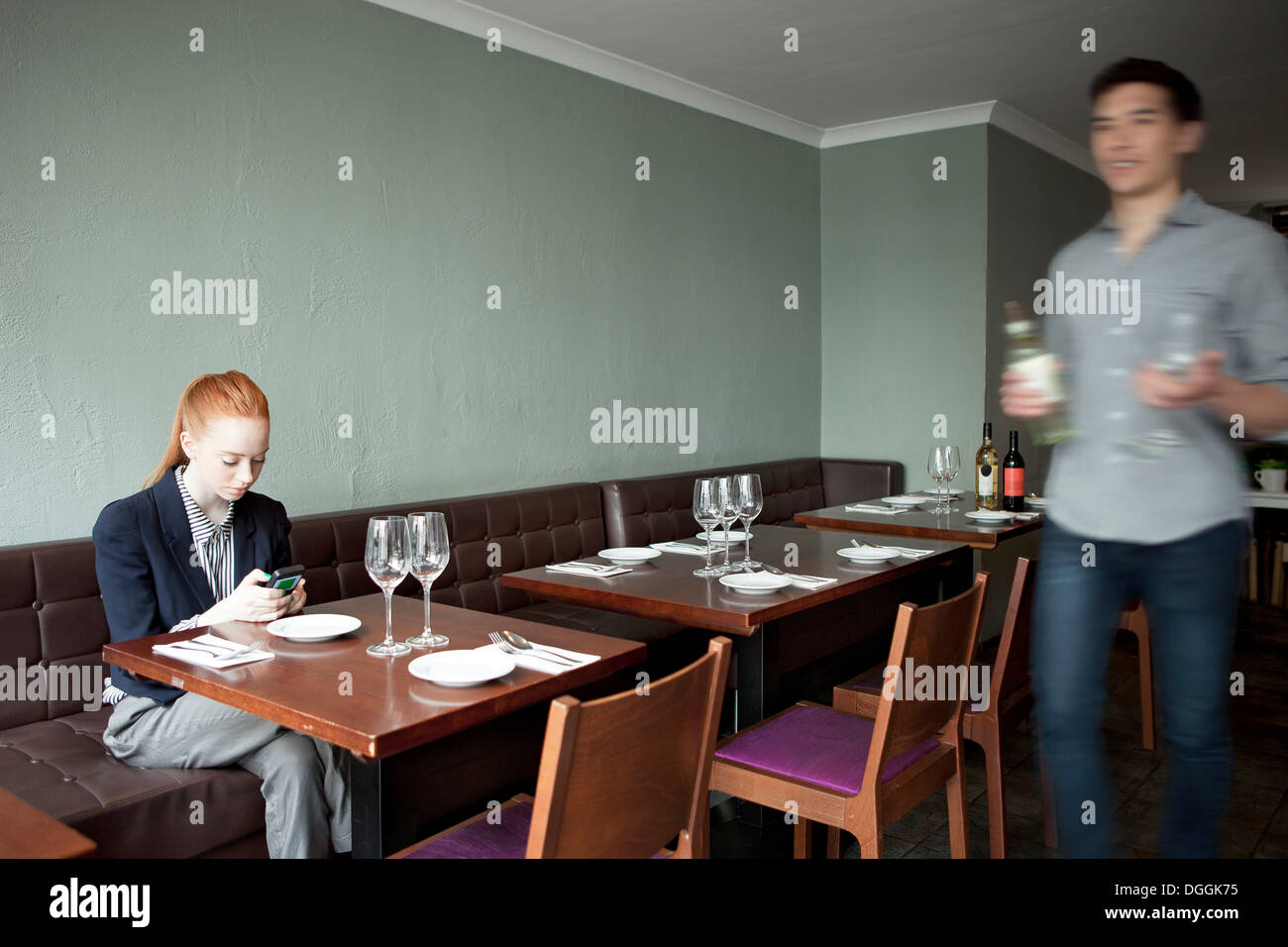 Young woman using cell phone in restaurant, serveur en passant devant Banque D'Images