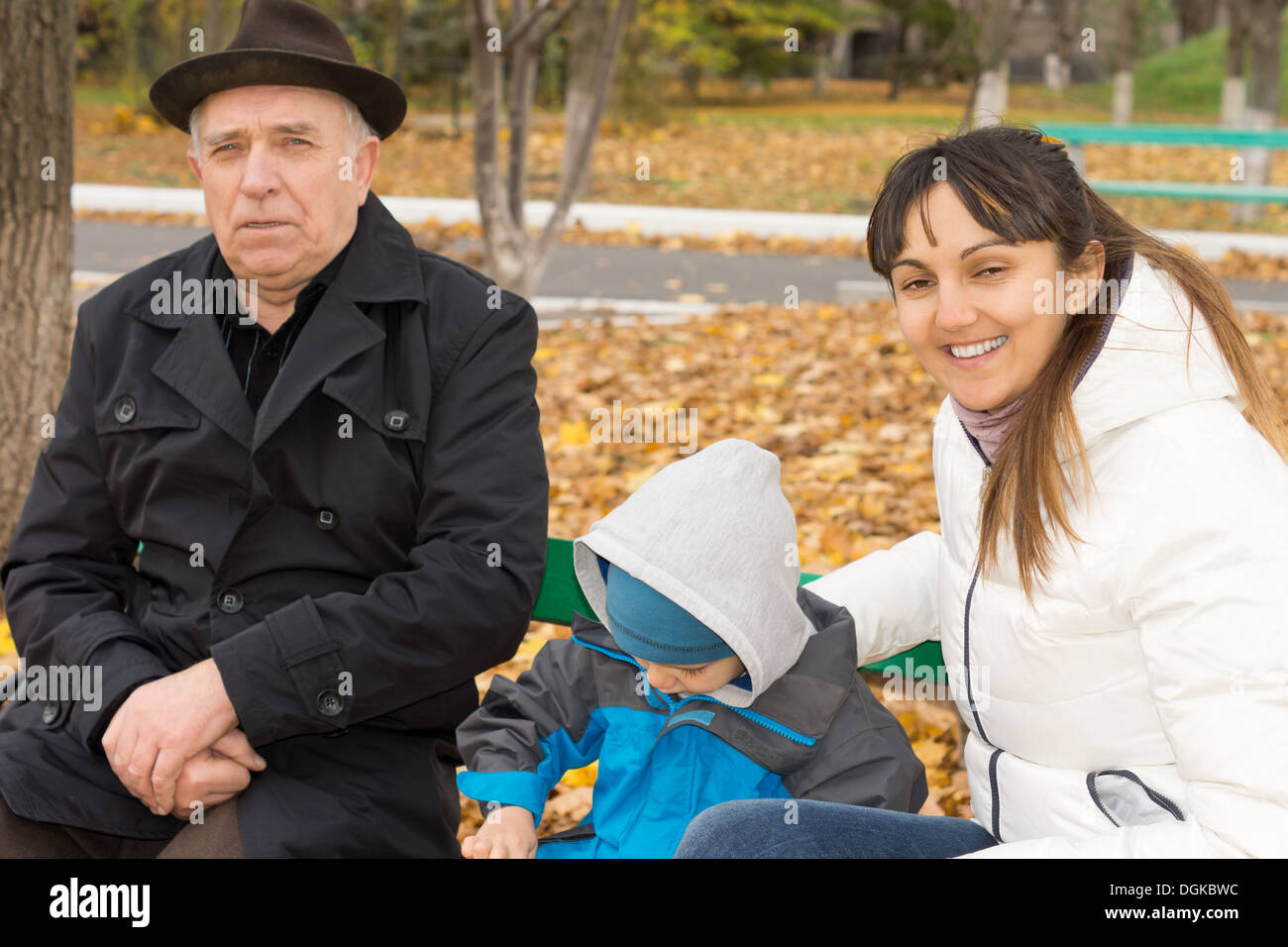Smiling friendly femme assise sur un banc de parc en bois avec son jeune fils et père âgé comme ils jouissent d'un jour d'automne dans l'air frais. Banque D'Images