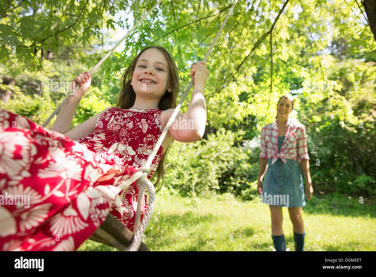 L'été. Une fille dans une robe d'été sur une balançoire sous un arbre feuillu. Une femme debout derrière elle. Banque D'Images