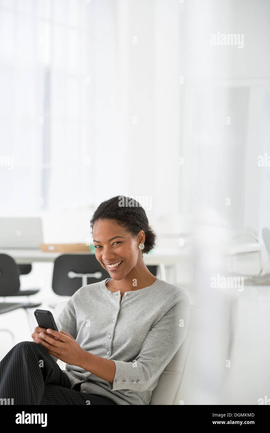 L'entreprise. Une femme assise dans une chaise confortable, contrôler son téléphone intelligent pour les messages. Banque D'Images L'entreprise. Une femme assise dans une chaise confortable, contrôler son téléphone intelligent pour les messages. Banque D'Images
