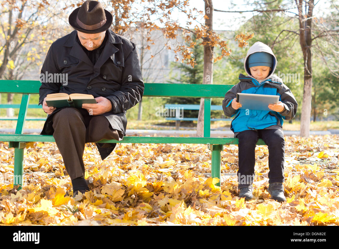 Homme âgé la lecture de son livre et un petit garçon à la recherche de sa tablette ordinateur reste ignorant les uns les autres à chaque extrémité le partage d'un banc de parc par une froide journée d'automne. Banque D'Images