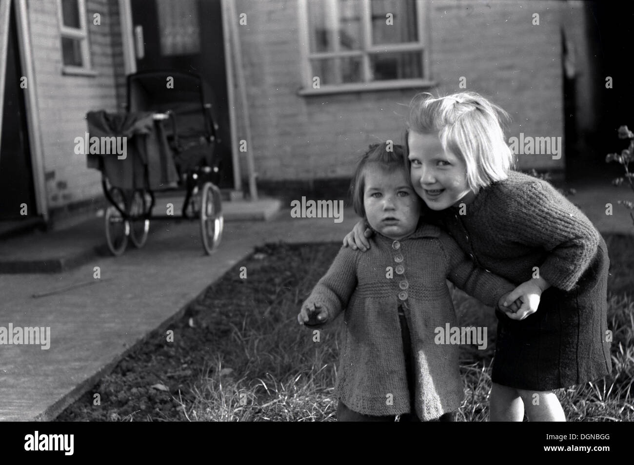 Image historique,1950 de deux petites filles dans un jardin à l'arrière, d'un aîné maintenant le plus petit de se tenir debout. Ancien style de la pram en arrière-plan. Banque D'Images