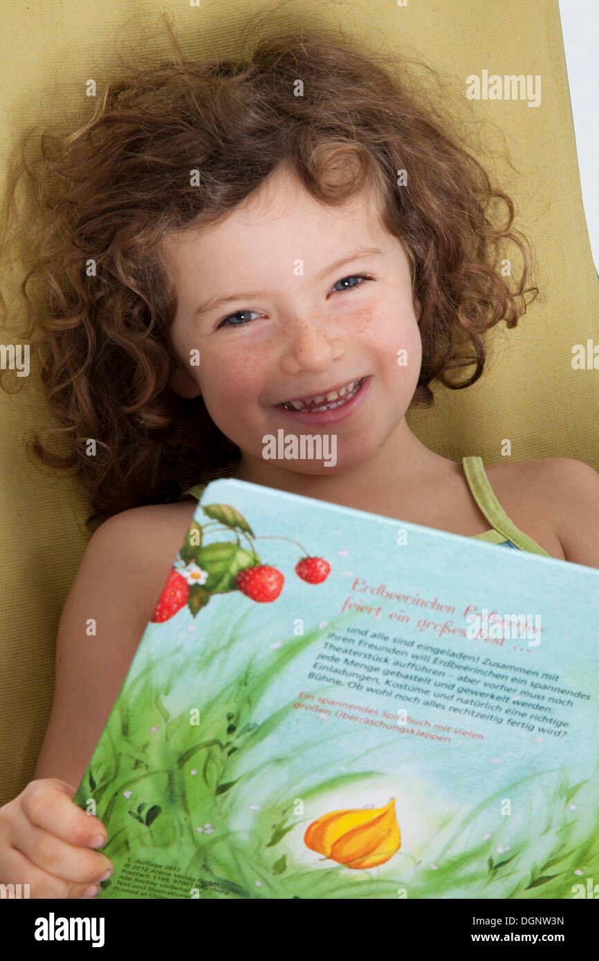 Fille aux cheveux roux, 5 ans, assis sur une chaise longue avec un livre pour enfants Banque D'Images