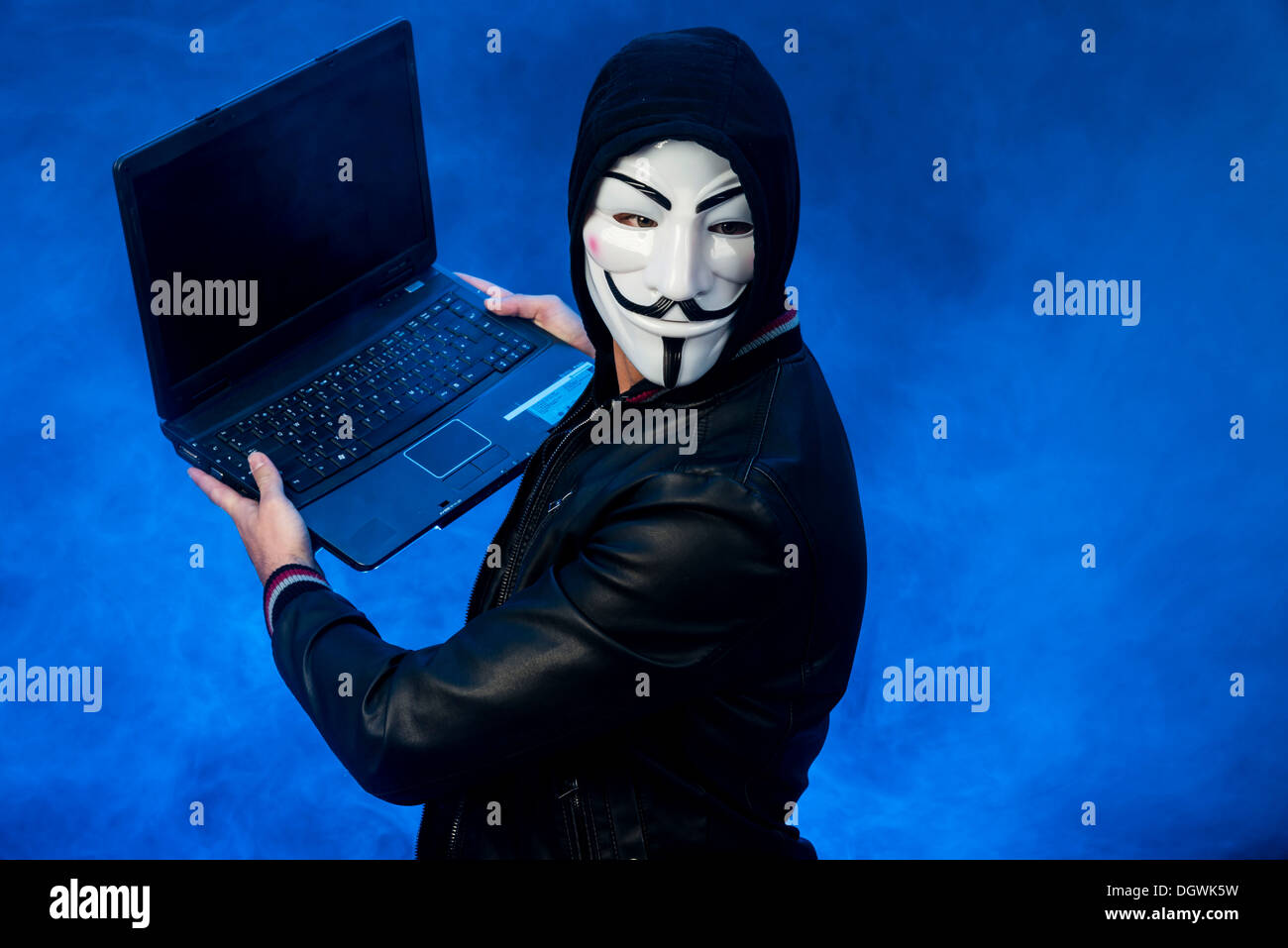 Jeune homme portant un masque anonyme avec un capuchon et une veste en cuir noire, tenant un ordinateur portable Banque D'Images